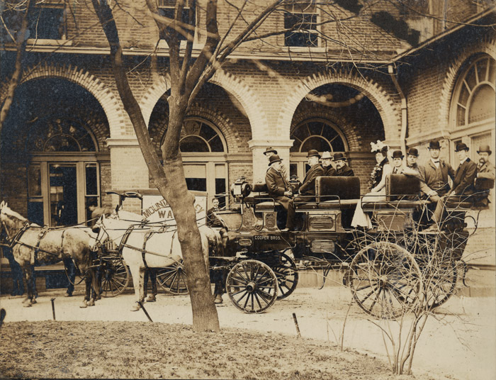 Horse drawn wagon with men and women in front of building with arches