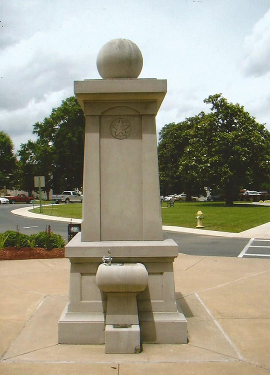 Stone monument with stone sphere on top and water fountain on its base with driveway and trees behind it