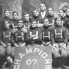 Group of young white men in uniforms and coach in a suit and tie with "Champions 07" sign