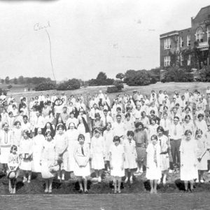Group of white men and women lined up in field with three-story building behind them
