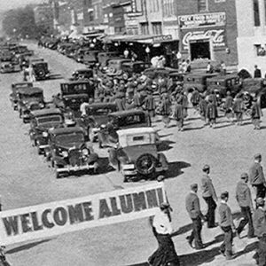 Parade with cars soldiers and floats on street with "Welcome Alumni" banner