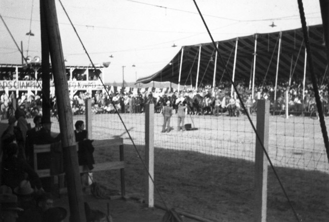 men and horses in outdoor arena with covered stands behind fencing as seen from opposite stands
