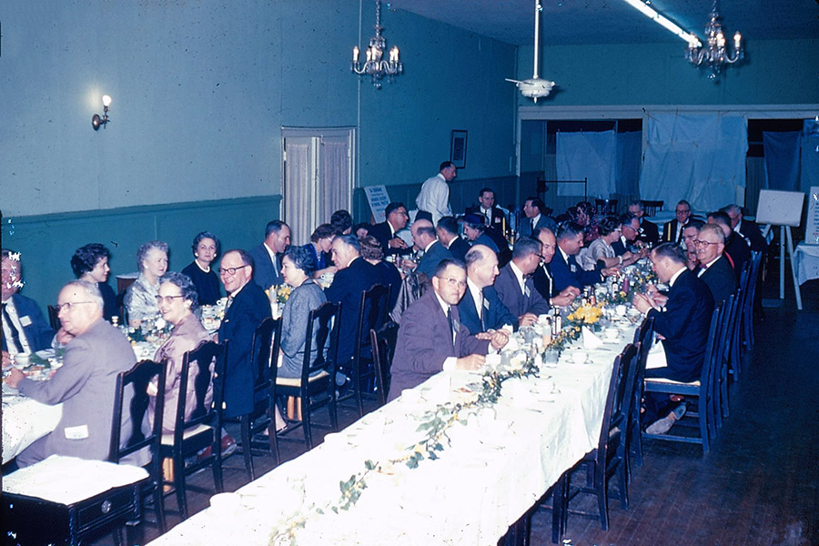 Group of older white men and women with glasses in formal clothing eating at long tables in dining hall