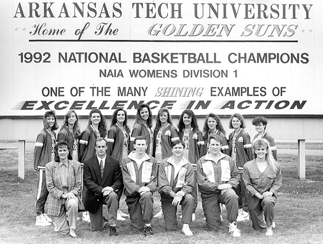 Group of young white women in matching athletic jackets with their male and female coaches kneeling in front of Arkansas Tech University billboard
