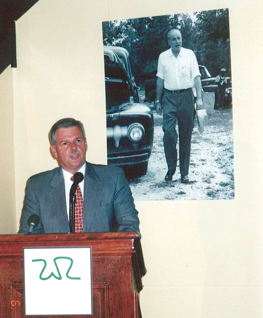 White man in suit speaking "W.R." logo lectern by portrait of white man with automobile