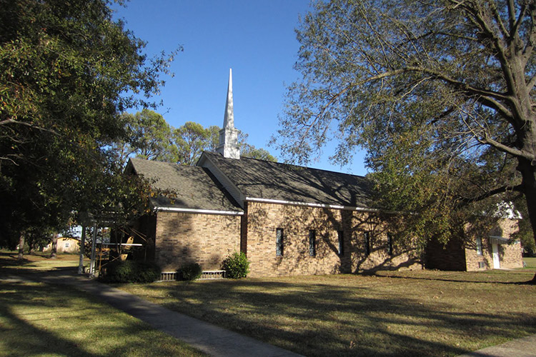 Side view of single-story brick church building with covered porch and steeple with tall tree next to it