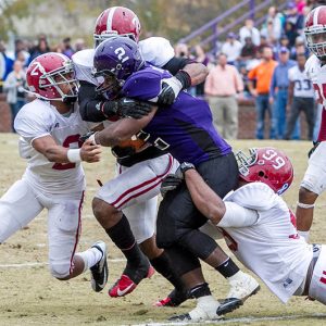Group of football players in red and white tackling a player in purple with crowded stands behind them