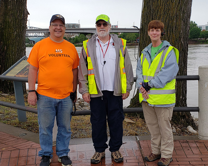 Two white men and woman in reflective vest standing by river with concrete bridge and city buildings in the background