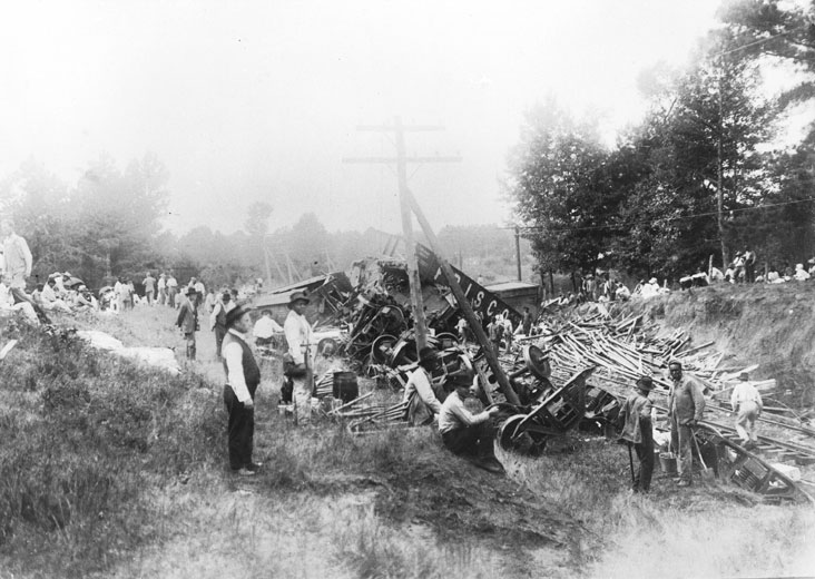 Mixed spectators gathered around wrecked train cars and their spilled cargo with trees in the background