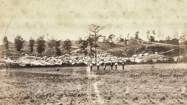 Group of men standing in field with rows of tents behind them