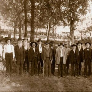 Group of white men wearing suits and hats standing in fenced-in field with trees and carriage behind them