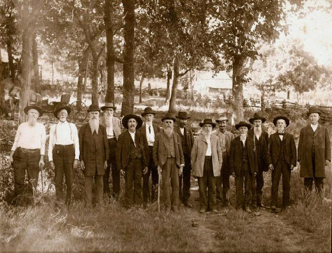 Group of white men wearing suits and hats standing in fenced-in field with trees and carriage behind them
