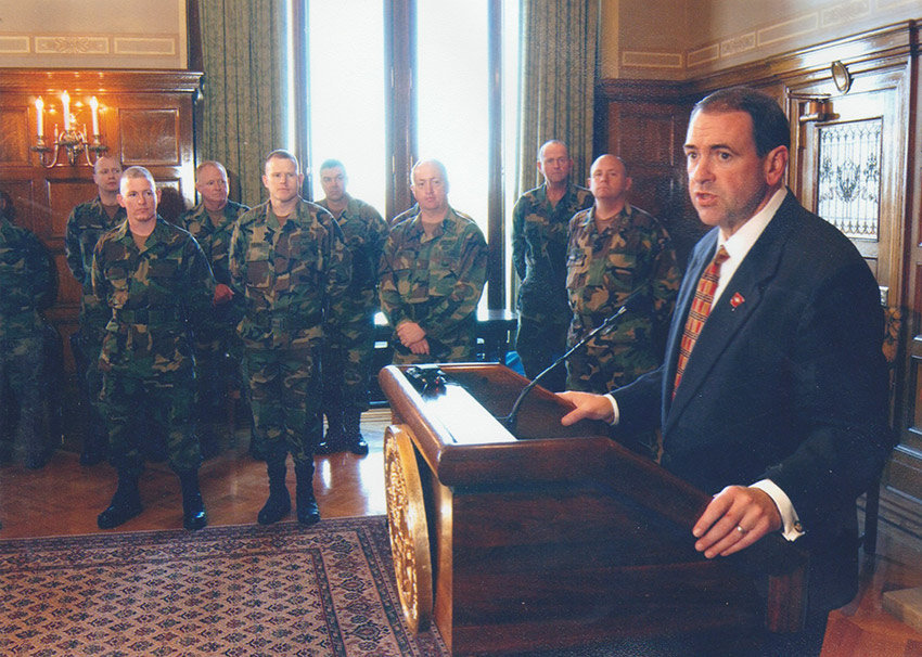 White man at lectern speaking in room alongside several people in camouflage