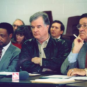 African-American man in suit sitting at table with two white men with crowd seated behind them