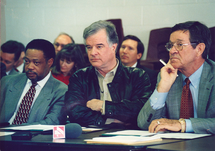 African-American man in suit sitting at table with two white men with crowd seated behind them