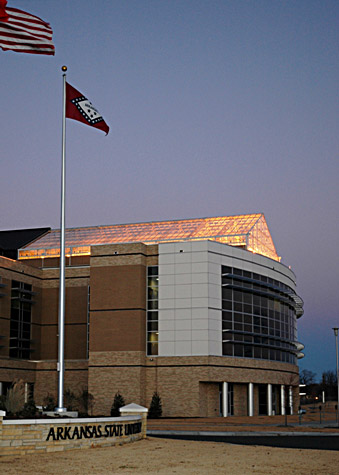multistory brick building with glass ceiling with lights showing through and two flagpoles outside with American and Arkansas flags