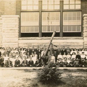 large group of people posing with large telescope in front of multistory stone and brick building