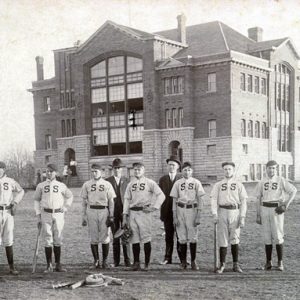 white baseball team poses in field in front of multistory stone and brick building