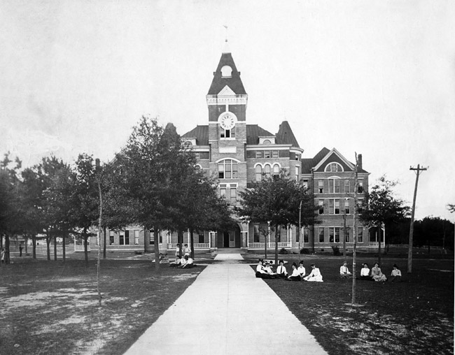 Students seated in school uniforms on quad outside large multistory brick building with clock tower