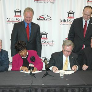 African-American woman and white men in suits sitting at table signing documents while white men in suits stand behind them
