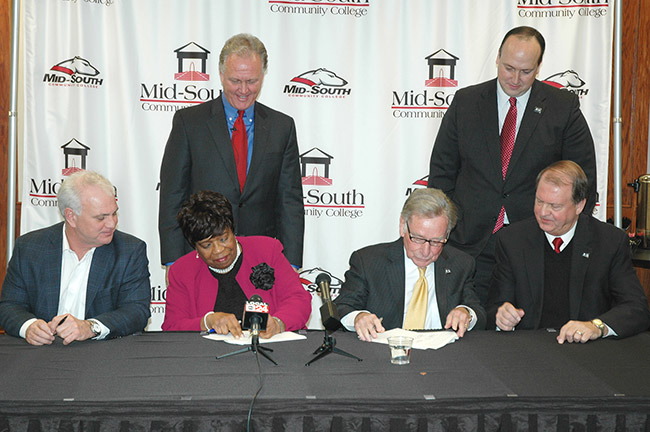 African-American woman and white men in suits sitting at table signing documents while white men in suits stand behind them