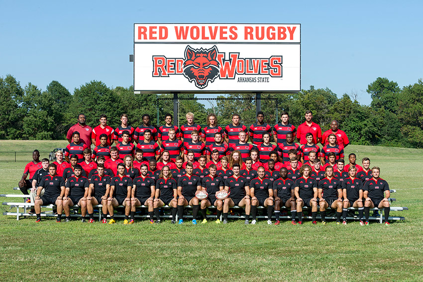 Group of young men and coaches in red and black  uniforms in front of "Red Wolves Rugby" sign