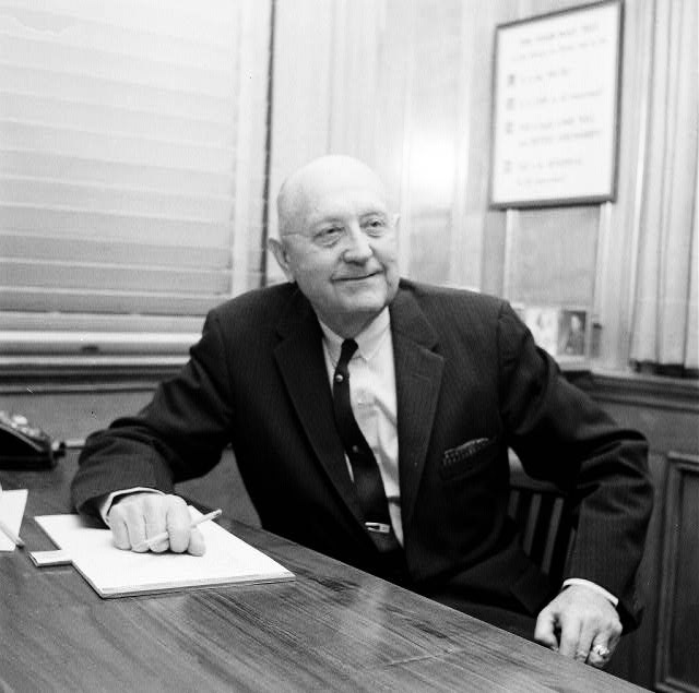 white man in suit and tie sitting at desk with pen and paper