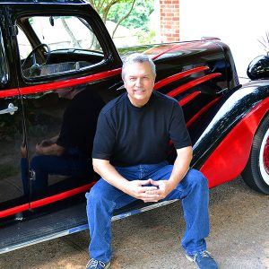 White man smiling sitting in driveway with black car with red accents on it