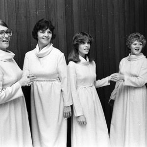 Four smiling white women in long white dresses with long sleeves and cowl necks