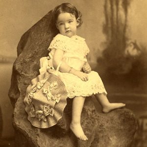 White female toddler in dress with hat sitting on a rock