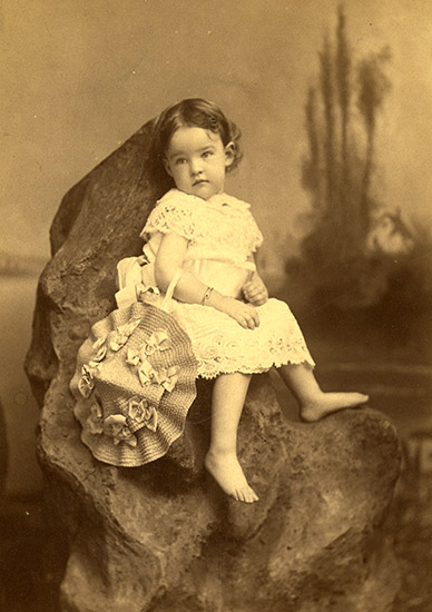 White female toddler in dress with hat sitting on a rock