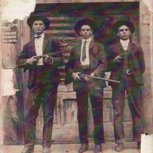 Three men in suits and hats with two holding shotguns and one holding a pistol