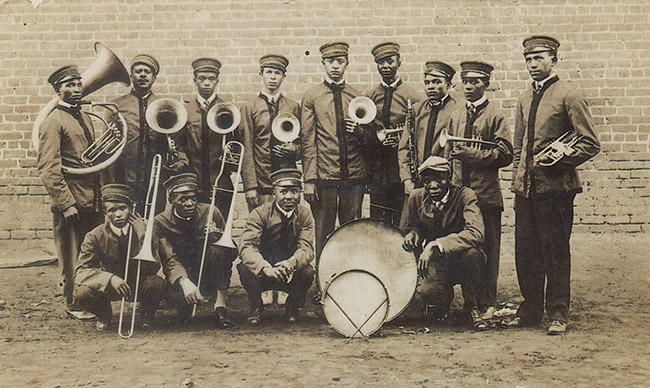 African-American brass band in uniforms with horns and drums posing outside brick building