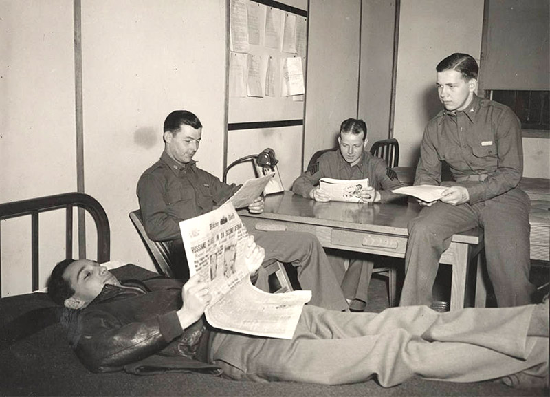 Four young men in military uniforms at rest in room with table, bed