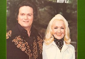 white man in embroidered black shirt and white woman in turtleneck and white jacket smiling in front of trees "Tony and Susan Alamo Tony Alamo sings Mister DJ"