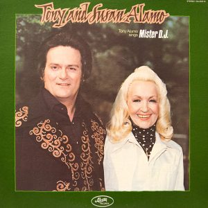 white man in embroidered black shirt and white woman in turtleneck and white jacket smiling in front of trees "Tony and Susan Alamo Tony Alamo sings Mister DJ"