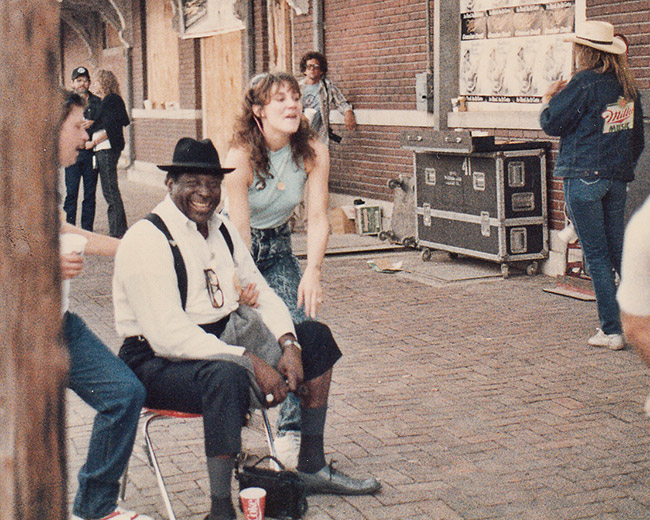African-American man in white shirt suspenders and hat sitting and laughing with white women posing with him outside brick building