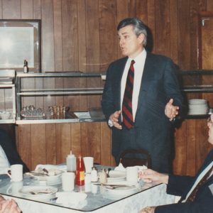 Older white men seated at table speaking to white man standing in suit in restaurant