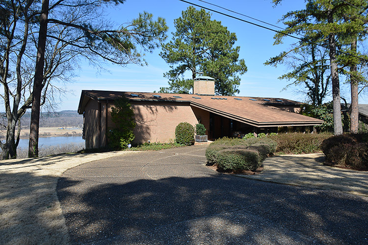 Single-story house with angled flat roof and covered entrance with chimney and river in the background