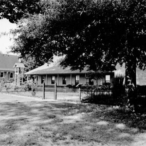 Single-story building with covered porch inside iron fence next to church building and trees in foreground