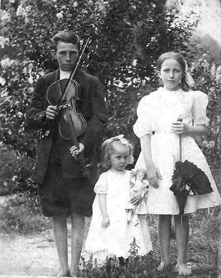White boy with fiddle standing next to white girl with umbrella and younger white girl with doll