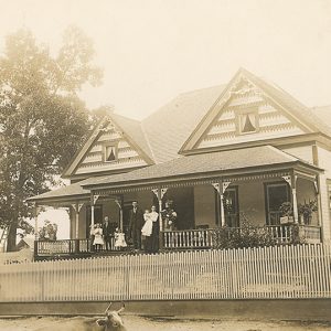 shingled house with two gables and ornate front porch with people in late early twentieth century style clothes