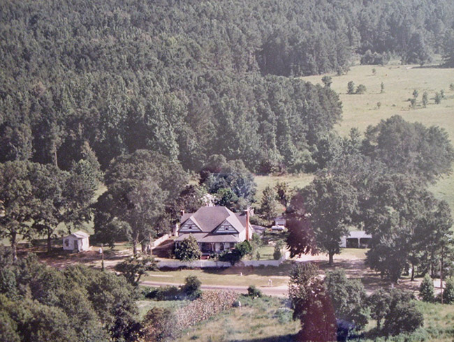 Multistory house and outbuildings on dirt road surrounded by tree covered countryside view from above