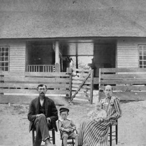 white family of four wearing early twentieth century attire sit in front of wooden house with wooden fence and gate