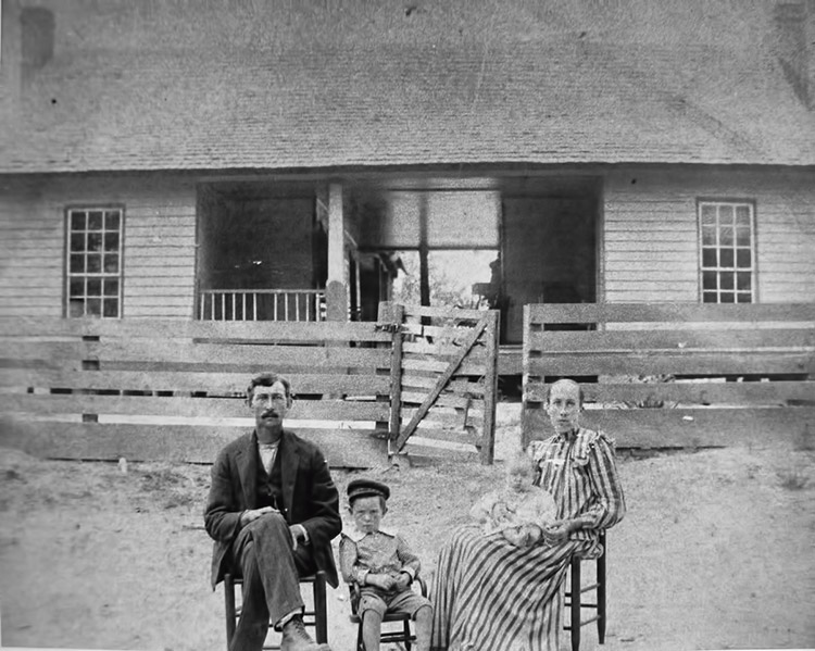 white family of four wearing early twentieth century attire sit in front of wooden house with wooden fence and gate