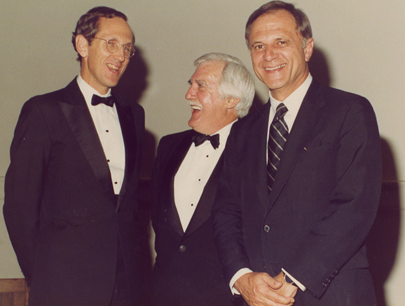White man in suit and tie smiling while white men in suit with glasses and older white man in suit laugh behind him