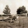 White man with hat standing with airplane in field with trees and house in the background