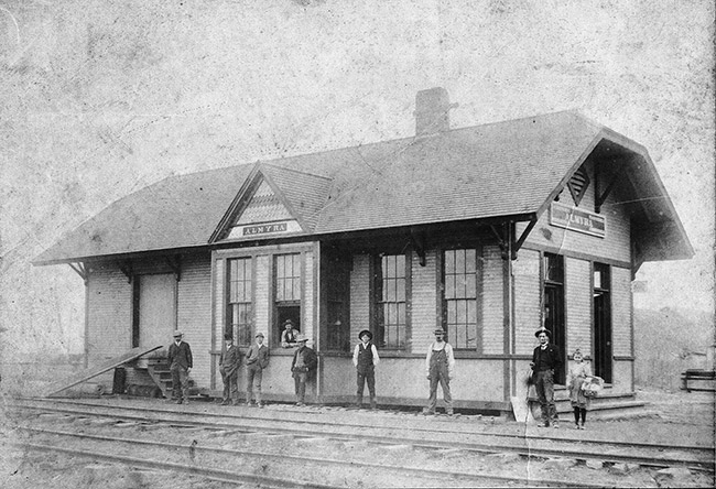 Eight white men with hats and one child at railroad depot building with train tracks