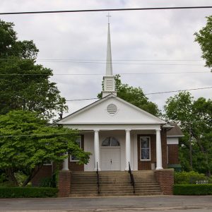 Front view of single-story brick church building with steeple and steps with iron railings and decorative tree on its left side