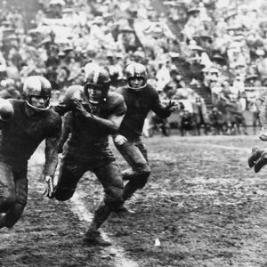 football players running down field covered in mud and onlookers filling the stands in background